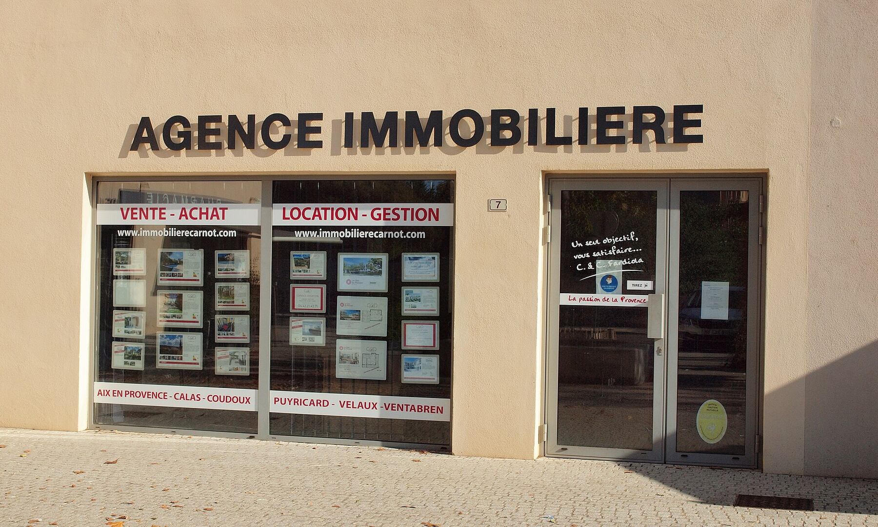 Façade d'une agence immobilière indépendante, avec l'enseigne « Agence immobilière » et des annonces affichées en vitrine