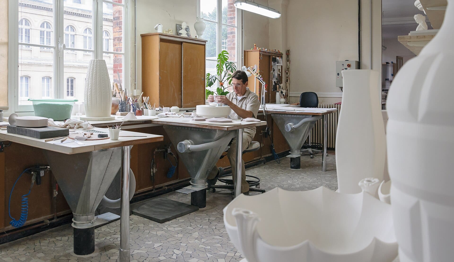 Atelier lumineux de porcelaine : un artisan travaille à son établi, pièces blanches au premier plan